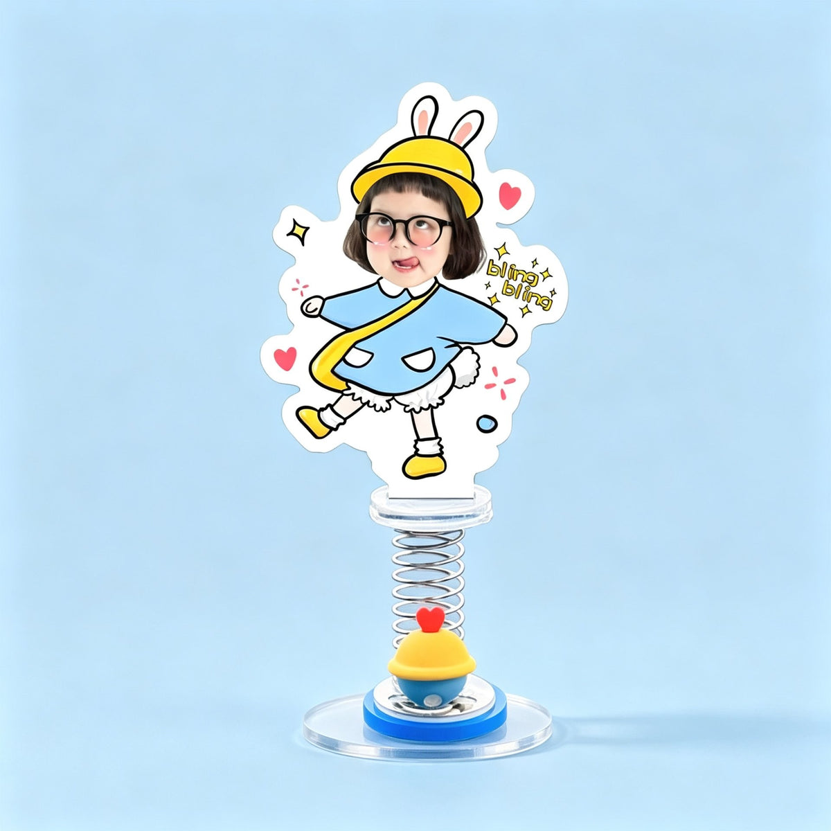Custom bunny hat kid photo spring bobblehead — cheeky kid in a yellow bunny hat and blue poncho strutting with bling bling stars, acrylic figure bouncing on a spring base